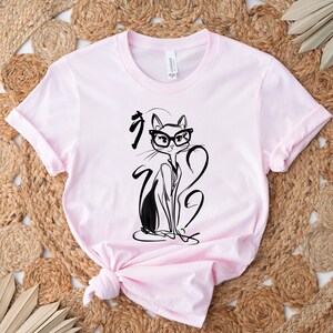 May include: Light pink t-shirt featuring a black and white illustration of a cat wearing glasses. The cat is drawn in a minimalist style with long, elegant lines. The shirt has a crew neck and short sleeves.