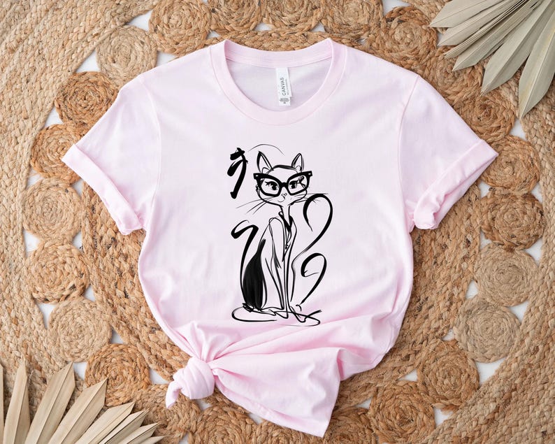 May include: Light pink t-shirt featuring a black and white illustration of a cat wearing glasses. The cat is drawn in a minimalist style with long, elegant lines. The shirt has a crew neck and short sleeves.