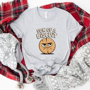May include: A light gray t-shirt featuring the text "SON OF A BISCUIT" above a cartoon biscuit with an angry face. The shirt is surrounded by a red and black plaid blanket, a gray and white patterned scarf, and a gray winter hat with pom-poms.
