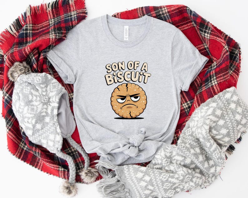 May include: A light gray t-shirt featuring the text "SON OF A BISCUIT" above a cartoon biscuit with an angry face. The shirt is surrounded by a red and black plaid blanket, a gray and white patterned scarf, and a gray winter hat with pom-poms.
