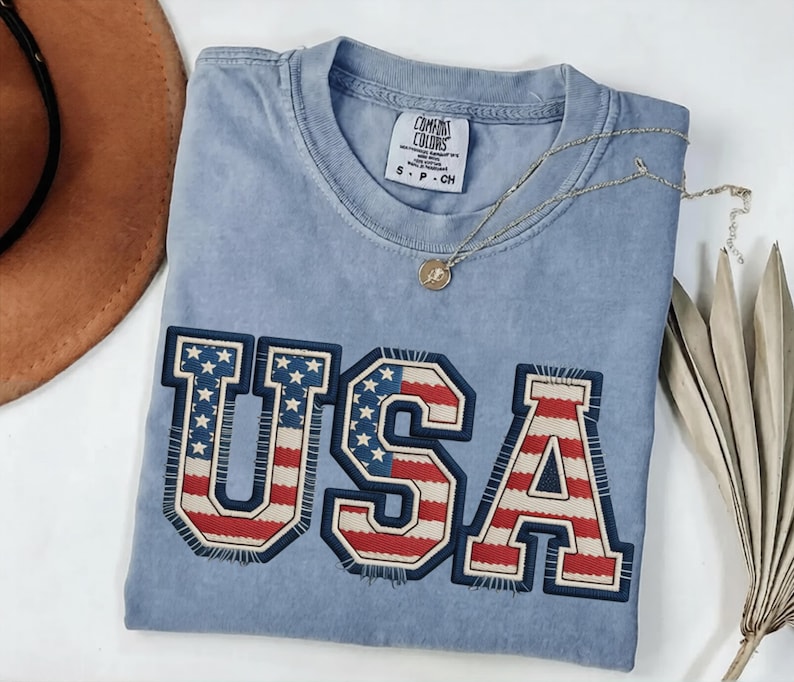 May include: A light blue Comfort Colors t-shirt with the word "USA" in a patriotic design. The letters are outlined in navy blue with a red, white, and blue flag pattern. A gold necklace and a brown hat are also visible.