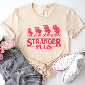 May include: A cream-colored t-shirt featuring a red graphic of four pugs riding bicycles, with the text "STRANGER PUGS" below. The shirt is paired with gray jeans and white sneakers.