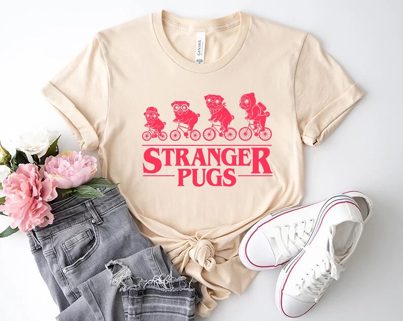 May include: A cream-colored t-shirt featuring a red graphic of four pugs riding bicycles, with the text "STRANGER PUGS" below. The shirt is paired with gray jeans and white sneakers.