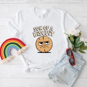 May include: White t-shirt with the text "SON OF A BISCUIT" above a cartoon cookie with an angry face. A rainbow and sunglasses are next to the shirt. The shirt is on a white wooden surface.