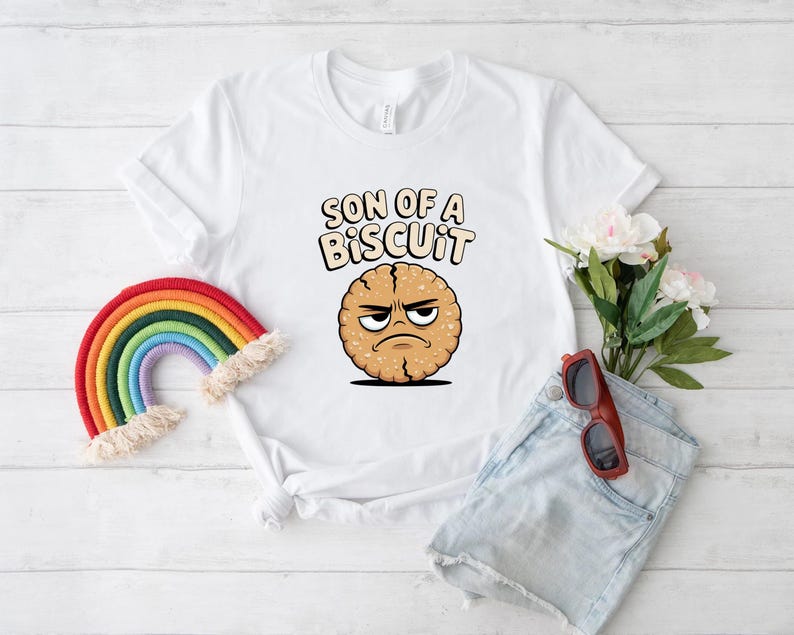 May include: White t-shirt with the text "SON OF A BISCUIT" above a cartoon cookie with an angry face. A rainbow and sunglasses are next to the shirt. The shirt is on a white wooden surface.