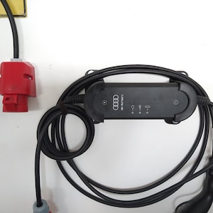 Könnte beinhalten: Ein schwarzes Ladekabel für Elektrofahrzeuge mit einem schwarzen Audi e-tron-Ladegerät, das an einer weißen Wand montiert ist. Das Kabel ist aufgewickelt und in eine rote Steckdose gesteckt.
