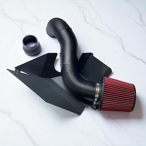 May include: Black automotive air intake system with a red air filter. The system includes a curved black pipe, a black shield, and a small black connector. The air filter is secured with a metal clamp.