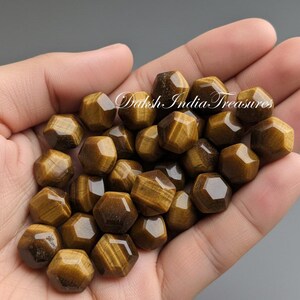 May include: A handful of faceted, golden-brown tiger's eye gemstone beads. The beads are hexagonal in shape and display a chatoyant effect. The text "Daksh India Treasures" is visible in the image.