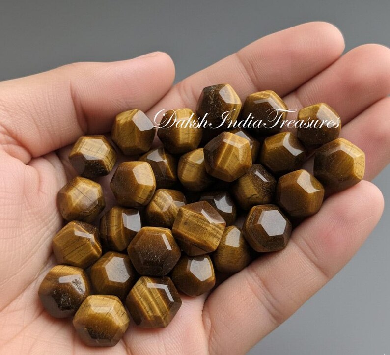 May include: A handful of faceted, golden-brown tiger's eye gemstone beads. The beads are hexagonal in shape and display a chatoyant effect. The text "Daksh India Treasures" is visible in the image.