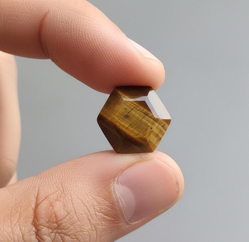 May include: A faceted, hexagonal tiger's eye gemstone held between two fingers. The gemstone has a warm, brown color with golden and dark brown stripes. The facets catch the light, creating a sparkling effect. The stone is approximately 0.75 inches in diameter.