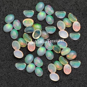 Puede incluir: Una colección de cabujones de ópalo ovalados e iridiscentes en varios tonos de blanco, crema y verde claro. Las piedras están dispuestas sobre una superficie gris oscuro.