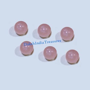 May include: Six pink rose quartz spheres, each approximately 2.5 cm in diameter, are arranged on a light blue background. The spheres are smooth and polished, with a slight sparkle.