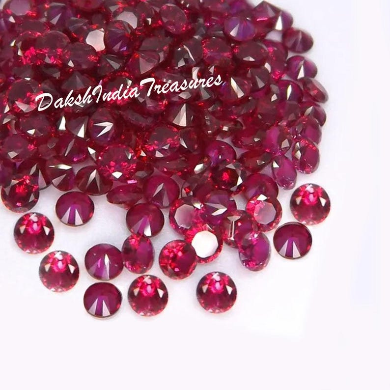 K&ouml;nnte beinhalten: Eine Ansammlung facettierter, runder, tiefroter Edelsteine. Die Edelsteine sind auf einer wei&szlig;en Oberfl&auml;che verstreut, mit dem Text "Daksh India Treasures" in kursiver Schrift. Die Edelsteine haben einen Brillantschliff.