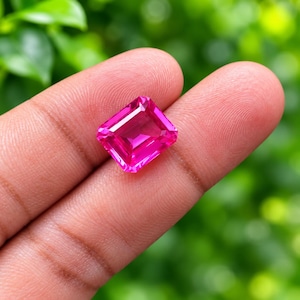 May include: A vibrant, hot pink gemstone cut into a rectangular emerald shape with faceted surfaces that reflect light. The stone displays a deep, saturated magenta hue and clear internal clarity, showcasing precise geometric edges and corners.