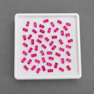 May include: A collection of small, rectangular, vibrant pink gemstones arranged in a white square display tray. Each stone features a faceted baguette cut, showcasing a consistent bright magenta hue and clear, reflective surfaces across the set.