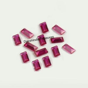 May include: A collection of thirteen loose, rectangular, baguette-cut gemstones in a vibrant magenta-pink hue. Each stone features faceted surfaces that reflect light. The text DakshIndiaTreasures is printed in black across the center of the stones.