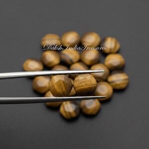 May include: Two faceted, round tiger's eye gemstones are held by silver tweezers, with a dark background. The gemstones have a golden-brown color with dark brown stripes. The background is filled with similar gemstones.