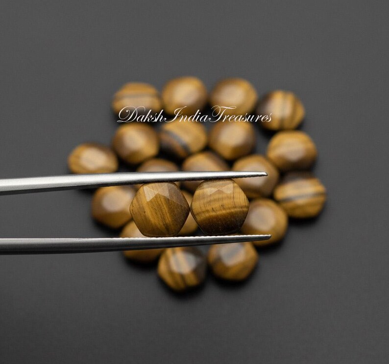 May include: Two faceted, round tiger's eye gemstones are held by silver tweezers, with a dark background. The gemstones have a golden-brown color with dark brown stripes. The background is filled with similar gemstones.