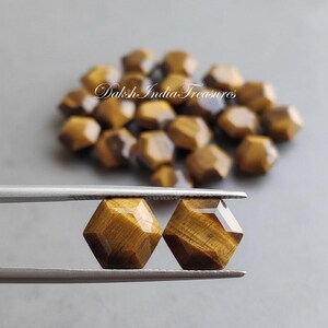 May include: Two faceted tiger's eye gemstones are held by tweezers, showcasing their warm brown and gold hues. A pile of similar gemstones is in the background. The image is on a gray surface, with the text "Daksh India Treasures" visible.