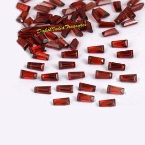 May include: A collection of rectangular, deep red gemstones scattered on a white surface. The gemstones have a polished, transparent appearance, with facets catching the light. The text "Daksh India Treasures" is visible in the upper left corner.