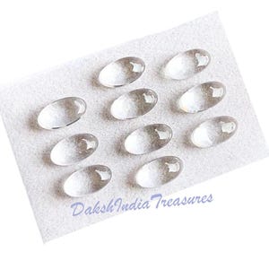 May include: Nine clear, oval glass cabochons are displayed on a white felt square. The cabochons have a slightly domed surface and a translucent appearance. The text "Daksh India Treasures" is printed in a light blue script.