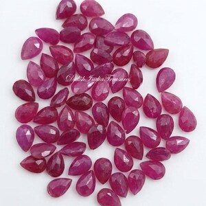 May include: A collection of loose, faceted, pear-shaped ruby gemstones in varying shades of deep pink and red. The stones are arranged in a cluster, and the text Daksh India Treasures is printed in white script across the center of the group.