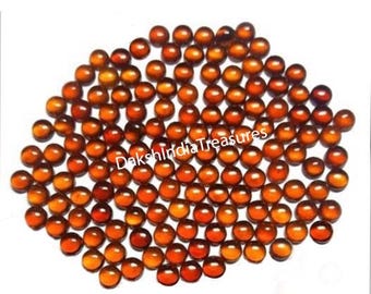 May include: A large pile of round, orange-brown cabochon gemstones. The gemstones are smooth and polished, and they are all about the same size.