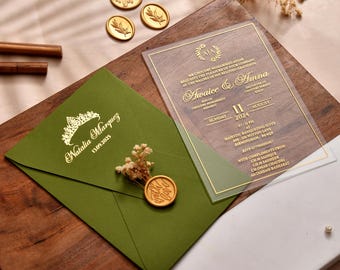 以下が含まれることがあります： 金色の文字と金のワックスシールが付いた、オリーブグリーンの結婚式の招待状の封筒。金色の文字が入った透明なアクリル製の招待状が重ねられています。ドライフラワーと追加のワックスシールも存在します。