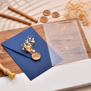 May include: A clear acrylic wedding invitation with gold lettering. The invitation features the names Samantha Pereira and Lorenzo Gamis, the date Tuesday, January 02, 2024, and the time 03:30 pm. The invitation also includes the location, Church of Our Lady of the Assumption, and the reception location, Dorchester Country Club, open bar.