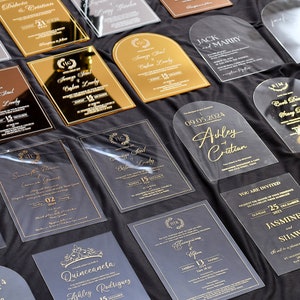 May include: A collection of clear acrylic wedding invitations with gold lettering. The invitations feature different designs, including arch shapes, floral wreaths, and calligraphy. The text on the invitations includes names, dates, and locations.