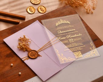 May include: A quinceañera invitation with gold text on a clear acrylic sheet, layered over a lilac envelope. The invitation includes the name "Reese Rosalinda" and event details. Decorative wax seals and dried flowers are also present.