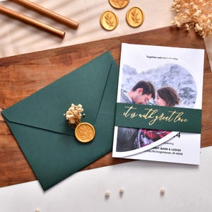 Può includere: Invito di nozze. Una busta verde scuro è sigillata con cera d'oro e fiori secchi. L'invito presenta una foto di una coppia e il testo "It is with great love". Sono visibili anche penne e sigilli d'oro.