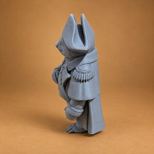 Puede incluir: Una figura gris impresa en 3D de un animal antropom&oacute;rfico vestido con un uniforme de estilo militar. El personaje lleva un sombrero bicornio alto, un abrigo largo con charreteras detalladas en los hombros y un cuello alto, visto de perfil.