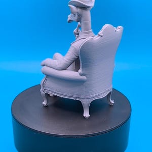 May include: A light gray 3D printed figurine of an anthropomorphic giraffe wearing a top hat and a suit, sitting in a detailed armchair. The figure is depicted in profile, holding a small pipe near its mouth, showcasing a smooth, matte finish.