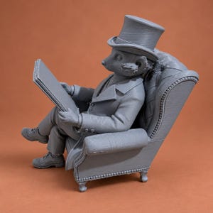 May include: A gray 3D printed figurine of an anthropomorphic meerkat wearing a top hat and a formal suit, sitting in a tufted armchair while holding an open book. The figure is depicted in a seated position with legs crossed, wearing casual shoes.
