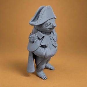 Puede incluir: Una figura gris impresa en 3D de un perezoso vestido con un uniforme de estilo militar. El perezoso lleva un sombrero bicornio, una chaqueta con charreteras en los hombros y una capa. La figura tiene un acabado mate liso y est&aacute; de perfil.