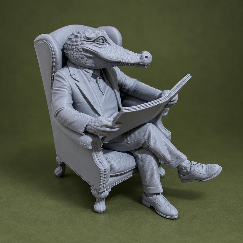 May include: A monochromatic gray figurine of an anthropomorphic alligator wearing a suit and tie, seated in a tufted wingback chair while holding an open newspaper. The figure features detailed scales on the head and hands, with formal shoes.
