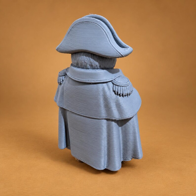 Puede incluir: Una figura impresa en 3D de color azul claro que lleva un sombrero bicornio y una capa larga con charreteras en los hombros. El objeto presenta un acabado mate uniforme y se muestra desde una perspectiva trasera, revelando su textura.