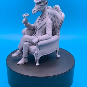 May include: A light gray 3D printed figurine of a giraffe with a human-like body wearing a formal suit, a bow tie, and a top hat. The giraffe is seated in a tufted armchair, holding a small mechanical device in one hand, with its legs crossed.