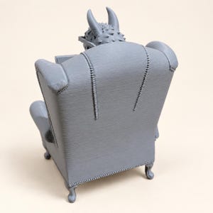 K&ouml;nnte beinhalten: Eine graue 3D-gedruckte Figur eines geh&ouml;rnten Wesens, das in einem Ohrensessel mit hoher R&uuml;ckenlehne sitzt. Der Sessel weist detaillierte Steppn&auml;hte entlang der R&uuml;ckenlehne auf und steht auf vier kleinen, geschwungenen Beinen.