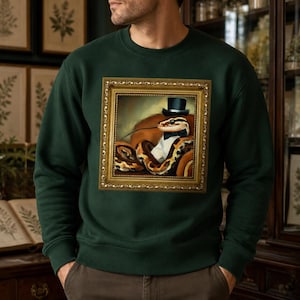 Op de afbeelding: Een donkergroene sweater met ronde hals en een centrale grafische print van een slang met een zwarte hoge hoed en een vlinderdas, afgebeeld in een goudkleurige, sierlijke fotolijst. De sweater heeft lange mouwen en een geribbelde hals.