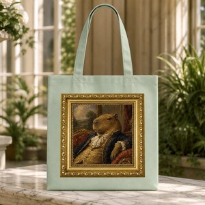 Puede incluir: Un bolso tote de color verde claro con un estampado rectangular de un capibara vestido con una casaca azul y un corbatín de encaje blanco del siglo XVIII, sentado en una silla de terciopelo rojo, dentro de un marco dorado detallado.