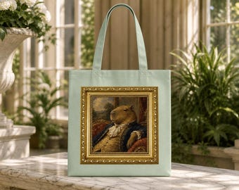 May include: A light green tote bag featuring a rectangular print of a capybara dressed in an 18th-century blue coat and white lace cravat, seated on a red velvet chair, all enclosed within an ornate, detailed gold-colored picture frame.