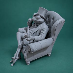 May include: A monochromatic gray figurine of a frog dressed in a formal suit, bow tie, and top hat, sitting in a tufted armchair while holding a smoking pipe. The frog has long, slender legs and is posed in a relaxed, seated position.