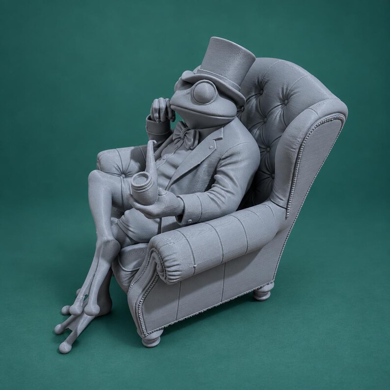 May include: A monochromatic gray figurine of a frog dressed in a formal suit, bow tie, and top hat, sitting in a tufted armchair while holding a smoking pipe. The frog has long, slender legs and is posed in a relaxed, seated position.