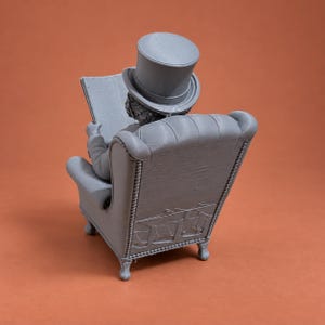 May include: A gray 3D printed figurine of a figure wearing a top hat, seated in a tufted armchair while holding an open book. The chair features detailed piping along the edges and carved legs, with a visible textured finish from the printing process.