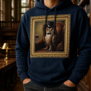 Könnte beinhalten: Ein marineblauer Kapuzenpullover mit einem rechteckigen Grafikdruck einer Eule, die einen schwarzen Zylinder und eine Fliege trägt und in einem braunen Ledersessel mit einem Glas Rotwein sitzt, alles eingerahmt von einem dekorativen goldfarbenen Rand.