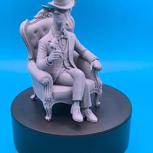 May include: A monochromatic gray 3D printed figurine of an anthropomorphic giraffe wearing a top hat, a formal suit with a vest, and a bow tie. The giraffe is seated in an ornate, tufted high-back armchair, holding a small hammer in one hand.