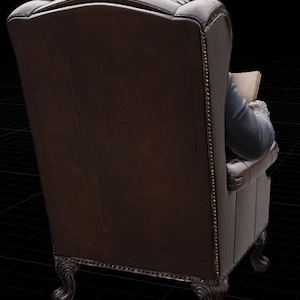 May include: A dark brown leather armchair with a high back and scrolled legs. The chair has a classic design with a tufted back and armrests. The back of the chair is visible, with a person in a blue jacket and white cuffs reading a book.