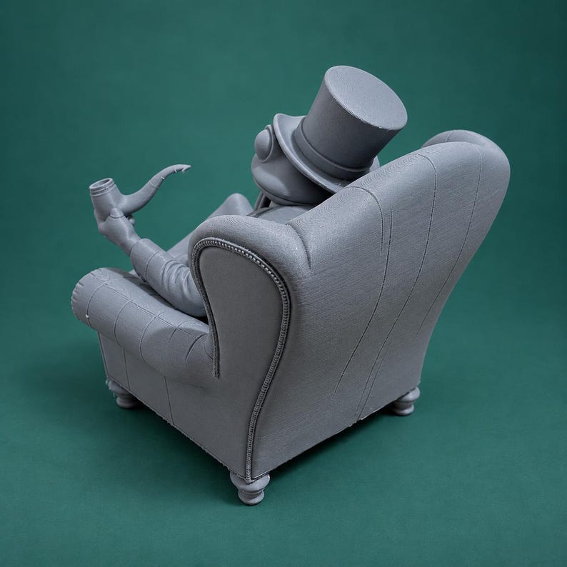 May include: A monochromatic gray figurine of a frog wearing a top hat, seated in a tufted armchair. The frog is holding a long-stemmed pipe in one hand. The sculpture features a smooth, matte finish and is depicted from a rear-facing angle.